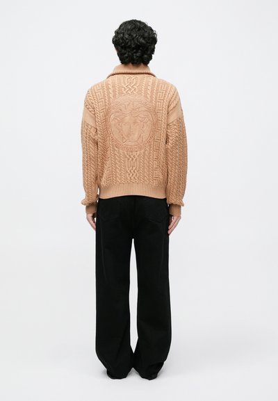 Persiku krāsas adīts džemperis ar riblietām aprocēm un apakšmalu, kas izceļ fakturētu rakstu un centrālu apaļu Medūzas emblemu; valkāts kopā ar melnām platām biksēm.