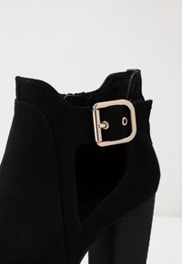 Zwarte suède enkellaars met blokhak en een gouden vierkante gesp aan de zijkant bij de uitsnijding.