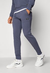 Námořnické modré tepláky s páskem, elastickými manžetami a bočními kapsami. Obsahují malou značkovou etiketu na boku. Hladká textura.