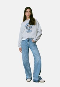 Mujer joven de pie con la mano en el bolsillo, vistiendo un sudadera gráfica gris claro, jeans de pierna ancha azul claro y zapatillas blancas.