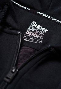 Sudadera negra con cremallera y etiqueta "Super Dry Sport", que presenta una textura de tela suave y una cremallera de nailon. La etiqueta indica múltiples estándares de tamaño.