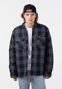 Black and gray checkered flannel shirt with two chest pockets, snap-button closure, and a relaxed fit. Worn over a white t-shirt.