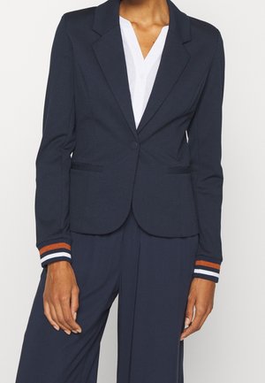 Personne portant un blazer bleu marine avec des poignets rayés orange et blanc sur une chemise blanche et un pantalon bleu marine assorti.