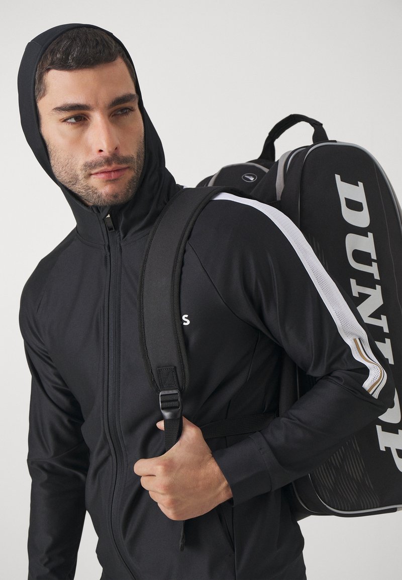 Schwarze, weite Sportjacke mit Reißverschluss und Kapuze, mit weißen Akzenten und Logo. Das Model trägt eine schwarze Sporttasche mit großer weißer Markenbeschriftung.