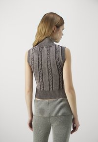 Top in maglione senza maniche e collo alto, di colore marrone, con motivo a trecce; orlo a coste. Indossato con pantaloni grigi a coste, che mostrano i dettagli della trama del tessuto.