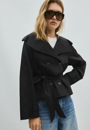 Femme portant une veste noire oversize, un crop top blanc, un short en jean longueur genou, des sandales noires à talons et de grandes lunettes de soleil foncées, debout devant un fond uni.
