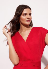 Czerwona sukienka kopertowa z krótkimi rękawami, dekoltem w szpic oraz widocznymi guzikami w talii; gładka faktura i jasny kolor podkreślają jej wygląd.