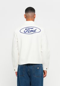 Man wearing white jacket with blue Ford logo on back and blue jeans, facing away against plain white background.