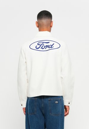 Mand iført hvid jakke med blåt Ford-logo på ryggen og blå jeans, vendt væk mod en ensfarvet hvid baggrund.