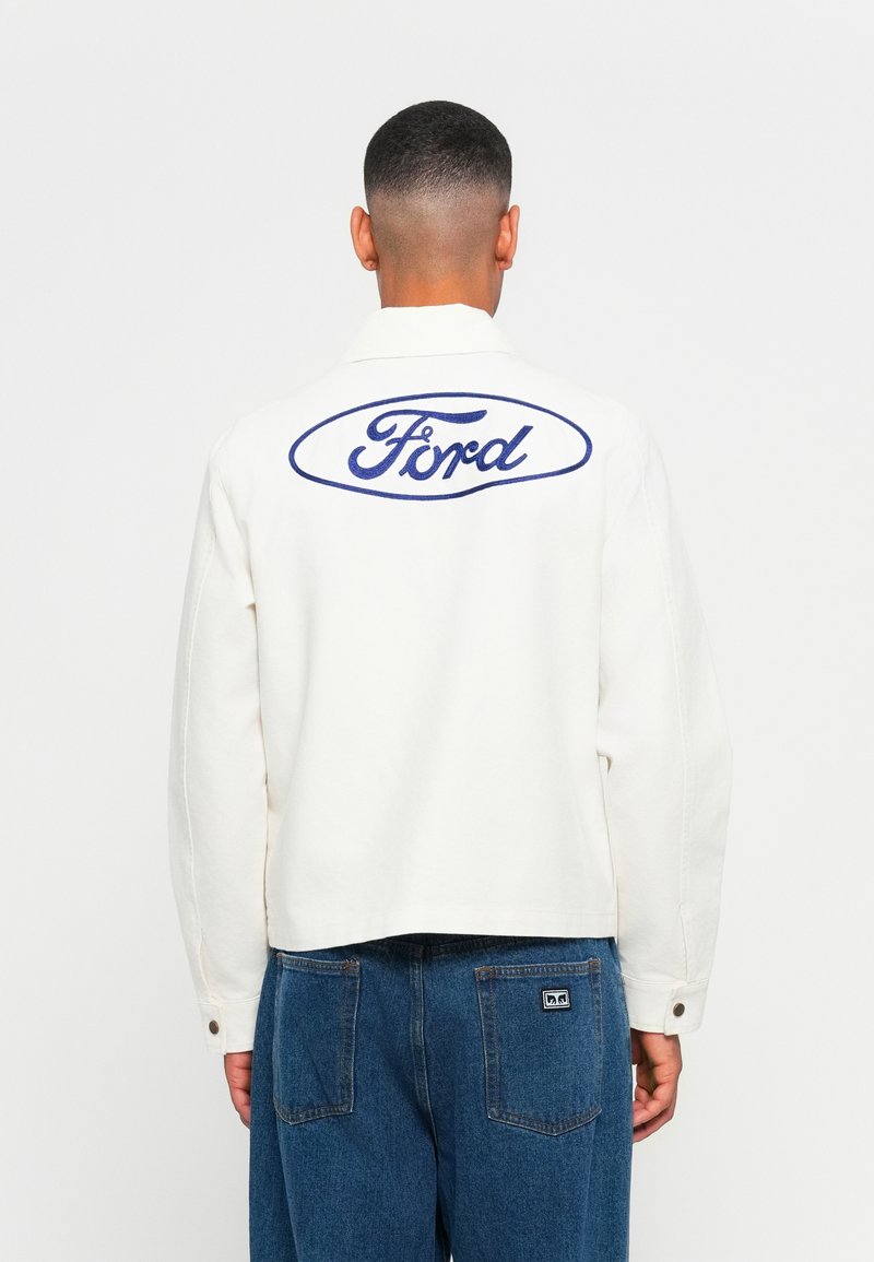 Man wearing white jacket with blue Ford logo on back and blue jeans, facing away against plain white background.