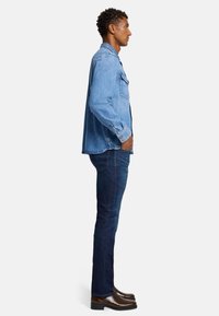 Lichtblauwe spijkerjas, voorzien van twee voorzakken, knoopsluitingen en een getailleerd ontwerp. Gecombineerd met donkerblauwe jeans en bruine schoenen.