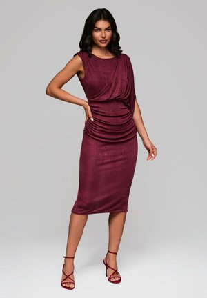 Vestido midi burdeos con tejido drapeado, detalles de fruncido y una silueta ajustada. Combinado con sandalias de tacón alto con tiras.