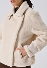Giacca in pile di colore crema, con texture, ampio collare, tasche con zip e dettaglio cintura con hardware dorato. Finitura in tessuto morbido.
