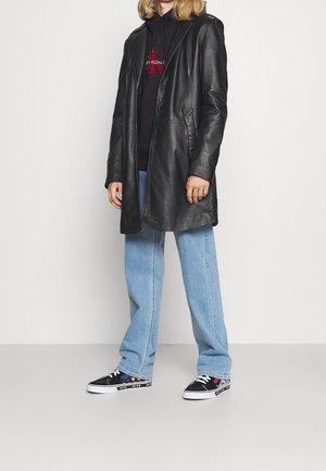 Cappotto lungo in pelle nera, jeans a gamba larga blu, sneakers nere con motivo. Il cappotto ha un cappuccio, tasche visibili e chiusura a bottoni.
