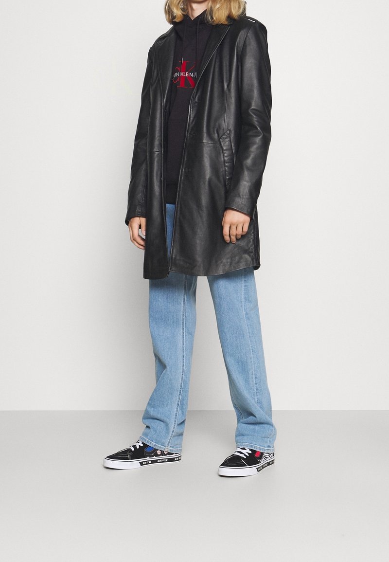 Cappotto lungo in pelle nera, jeans a gamba larga blu, sneakers nere con motivo. Il cappotto ha un cappuccio, tasche visibili e chiusura a bottoni.