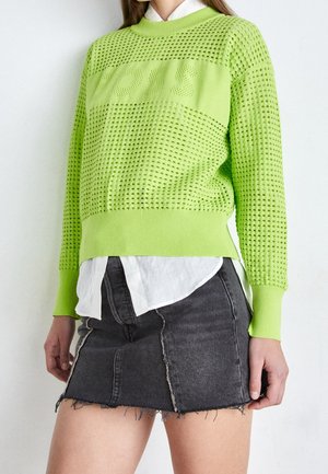 Pull en tricot vert citron avec une texture perforée, arborant le mot "BOSS" au centre, porté par-dessus une chemise blanche et une jupe en denim noire.