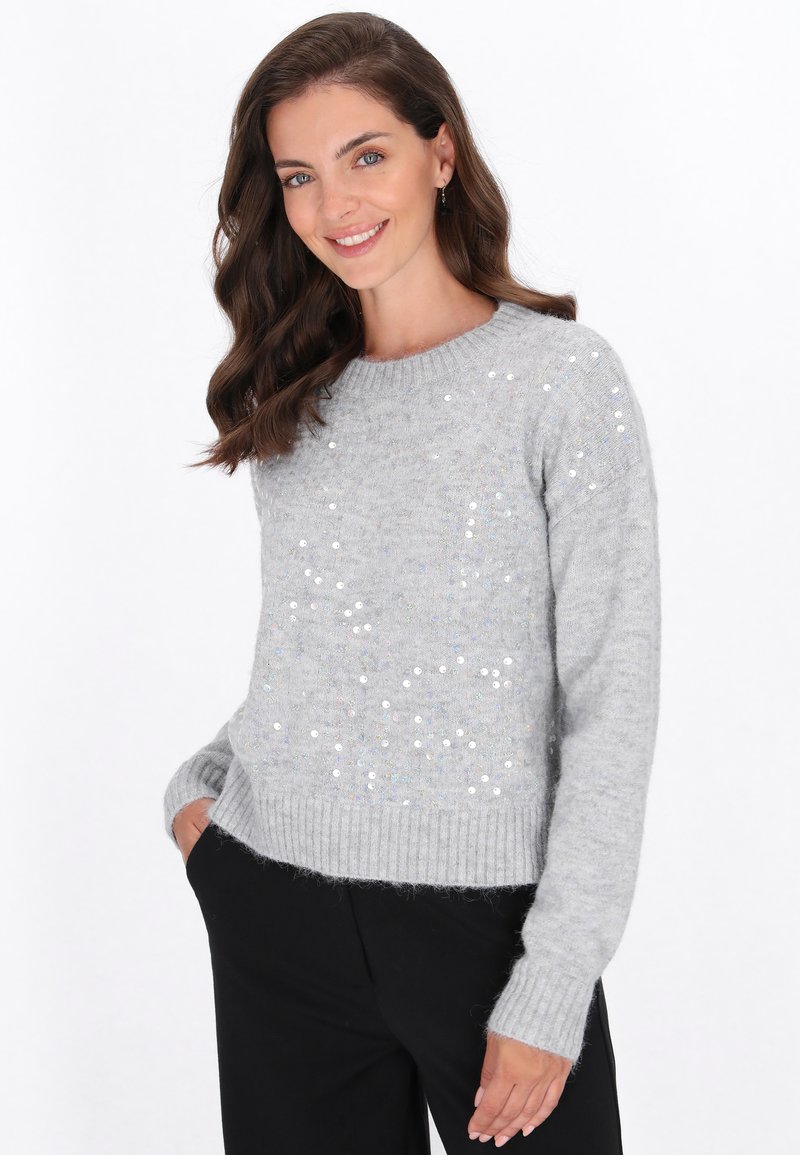Glunderende vrouw met golvend bruin haar, gekleed in een lichtgrijze pailletten trui en zwarte broek, met handen in de zakken tegen een witte achtergrond.