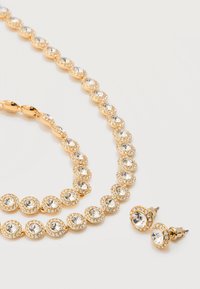 Κολιέ με χρυσό τόνο και σκουλαρίκια καρφάκια που ταιριάζουν, και τα δύο με στρογγυλά διαυγή κρύσταλλα περιβαλλόμενα από μικρότερα κρύσταλλα σε λευκό φόντο.