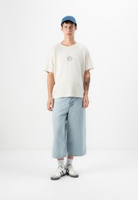 Beige short-sleeve t-shirt with circular logo, light blue wide-leg denim jeans, white sneakers with black stripes, and blue cap.