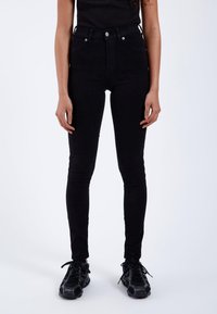 Femme portant un jean skinny noir taille haute et des chaussures de sport noires, debout devant un fond uni.