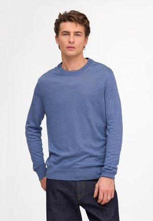 Jonge man met een blauwe trui met ronde hals en donkere spijkerbroek, staand met één hand in zijn zak tegen een effen witte achtergrond.