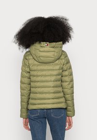 Doudoune vert olive avec une capuche ajustée, design matelassé horizontal et logo brodé au col. Associée à un jean bleu.