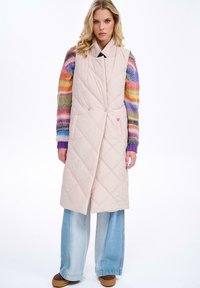 Pikowana długa kamizelka w jasnym beżu, z kołnierzem wykładanym, podwójnymi guzikami i luźnym krojem, zestawiona z wielokolorowym swetrem dzianinowym oraz szerokimi dżinsami.