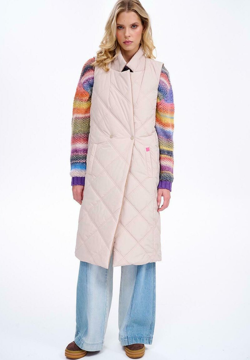 Pikowana długa kamizelka w jasnym beżu, z kołnierzem wykładanym, podwójnymi guzikami i luźnym krojem, zestawiona z wielokolorowym swetrem dzianinowym oraz szerokimi dżinsami.