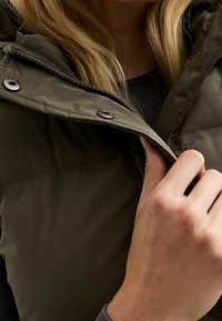 Vihreä oliivinvärinen puffa vest, jossa on vetoketjusulku, korkea kaulus ja metalliset yksityiskohdat. Vestissä on teksturoitu pinta ja sileä vuori.