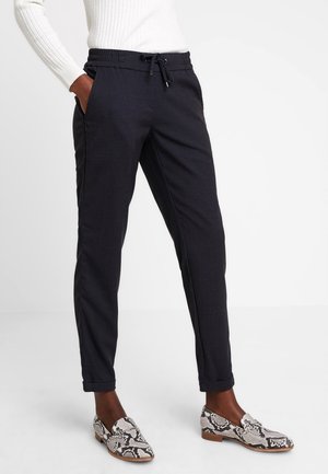 Personne portant un pantalon noir à cordon, un pull blanc côtelé et des mocassins à motif peau de serpent, debout les mains dans les poches.
