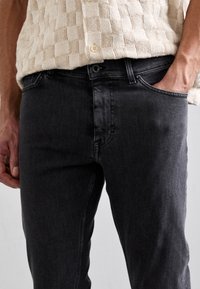 Mustat denimfarkut, joissa on kapea istuvuus, hienovaraisesti teksturoitu pinta ja perinteinen viiden taskun malli. Nappikiinnitys ja vetoketju näkyvissä.