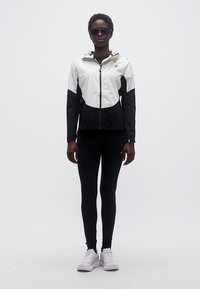 Ενήλικας που φοράει μαύρο και άσπρο μπουφάν με κουκούλα, μαύρο κολάν, άσπρα αθλητικά παπούτσια και ανακλαστικά γυαλιά ηλίου, στέκεται μπροστά σε μονόχρωμο φόντο.