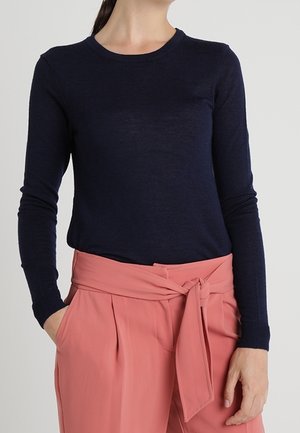 Femme portant une chemise bleu marine ajustée à manches longues, rentrée dans un pantalon taille haute couleur saumon avec ceinture nouée, les mains dans les poches.
