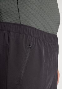 Schwarze Sportshorts mit elastischem Bund, ausgestattet mit einer seitlichen Reißverschlusstasche und über einem strukturierten grünen Oberteil getragen.