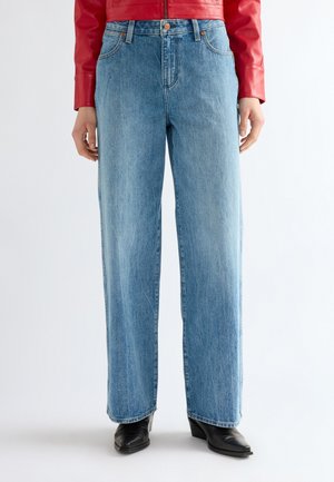 Persona che indossa jeans celesti a gamba larga, stivali neri con punta appuntita e una giacca rossa, in piedi davanti a uno sfondo semplice.