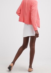 Giacca corallo con una vestibilità rilassata e tessuto texture, abbinata a una mini gonna bianca. Il modello indossa delle ballerine beige chiaro.