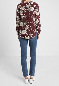 Blouse florale bordeaux à manches longues, ornée de fleurs blanches et vertes. Associée à un jean en denim bleu et des talons ouverts blancs.
