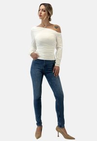 Witte off-the-shoulder top met gerimpelde textuur, gecombineerd met nauwsluitende blauwe jeans. Het model draagt beige hoge hakken. Eenvoudige achtergrond.