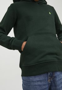 Personne portant un sweat à capuche vert foncé avec une poche avant et un logo vert clair, les mains dans la poche, assorti à un jean bleu en denim.