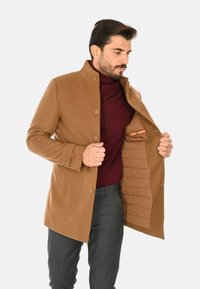 Cappotto in lana marrone, texturizzato, con colletto rialzato, chiusura a bottoni e fodera trapuntata visibile. Il modello mostra l'interno con tasca nascosta.