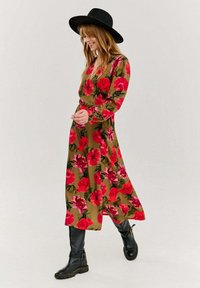 Kwiatowa sukienka maxi w oliwkowym kolorze, z dużymi czerwonymi i różowymi kwiatami. Długie rękawy i dopasowana talia, w połączeniu z czarnymi wysokimi botkami i kapeluszem o szerokim rondzie.