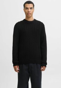 Jonge man die staat, gekleed in een zwart kabelgebreid trui en donkere broek, tegen een plain witte achtergrond.