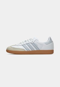 Baltikie snickers Adidas Samba ar gaišiem pelēkiem svītrojumiem, bēšīgu purngali un gumijas zoli, parādīti sānskatā uz balta fona.