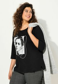 T-shirt nera con maniche a tre quarti caratterizzata da una stampa grafica di un volto. Abbinata a pantaloni grigi texturizzati e a un cardigan grigio drappeggiato sulla spalla.