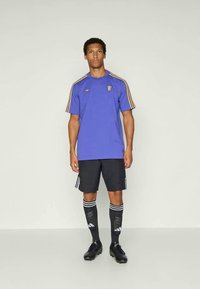 Άνδρας φοράει αθλητική μπλούζα μωβ με κοντά μανίκια και χρυσές ρίγες, μαύρες βερμούδες, μαύρες έως το γόνατο κάλτσες και μαύρα παπούτσια ποδοσφαίρου, σε απλό φόντο.