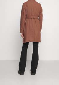 Abrigo de mezcla de lana marrón con cintura ceñida, que presenta un diseño entallado, mangas largas y una parte trasera recta, acompañado de pantalones negros de campana.