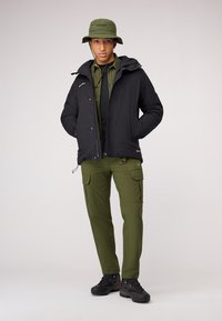 Giacca nera isolata con cappuccio, camicia utility verde oliva, pantaloni cargo e scarpe da trekking nere. Include un cappello a secchiello verde.