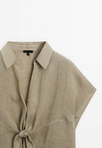 Chemise beige en lin à manches courtes avec col et nœud à nouer à l'avant, présentée sur fond blanc.