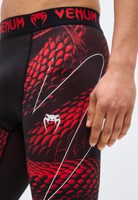 Κοντινό πλάνο ενός ατόμου που φοράει κολάν με μοτίβο από φολίδες φιδιού σε μαύρο και κόκκινο χρώμα, με τη ζώνη μέσης "VENUM" και λογότυπο φιδιού με δόντια κοντά στον μηρό.