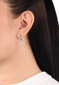 Acercamiento a la oreja de una mujer con cabello lacio y oscuro, que lleva un pequeño pendiente de aro de plata con un charm en forma de rosa.